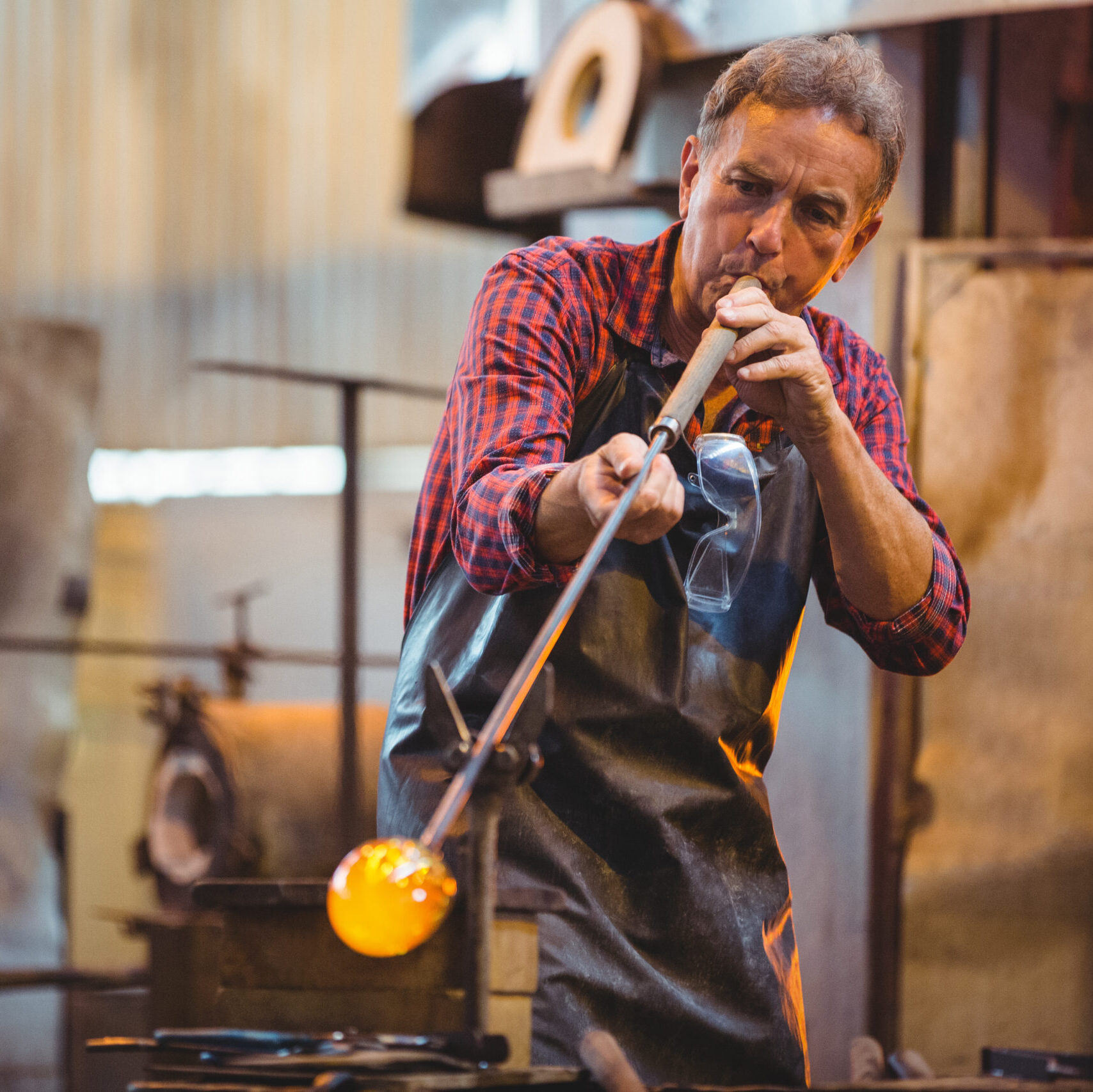 A glassblower shaping molten glass using a blowpipe in a workshop.
